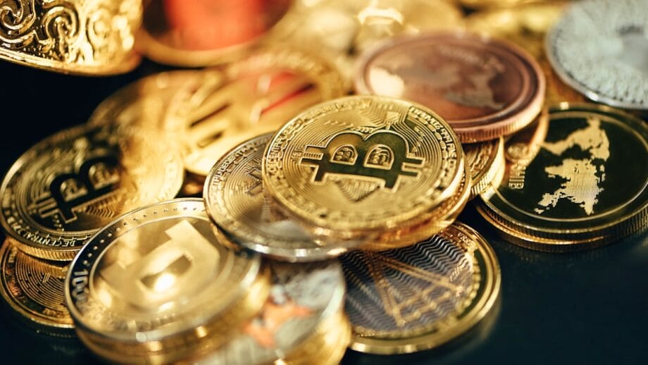 A pile of assorted cryptocurrency coins, including prominent Bitcoin symbols, displayed on a dark surface.