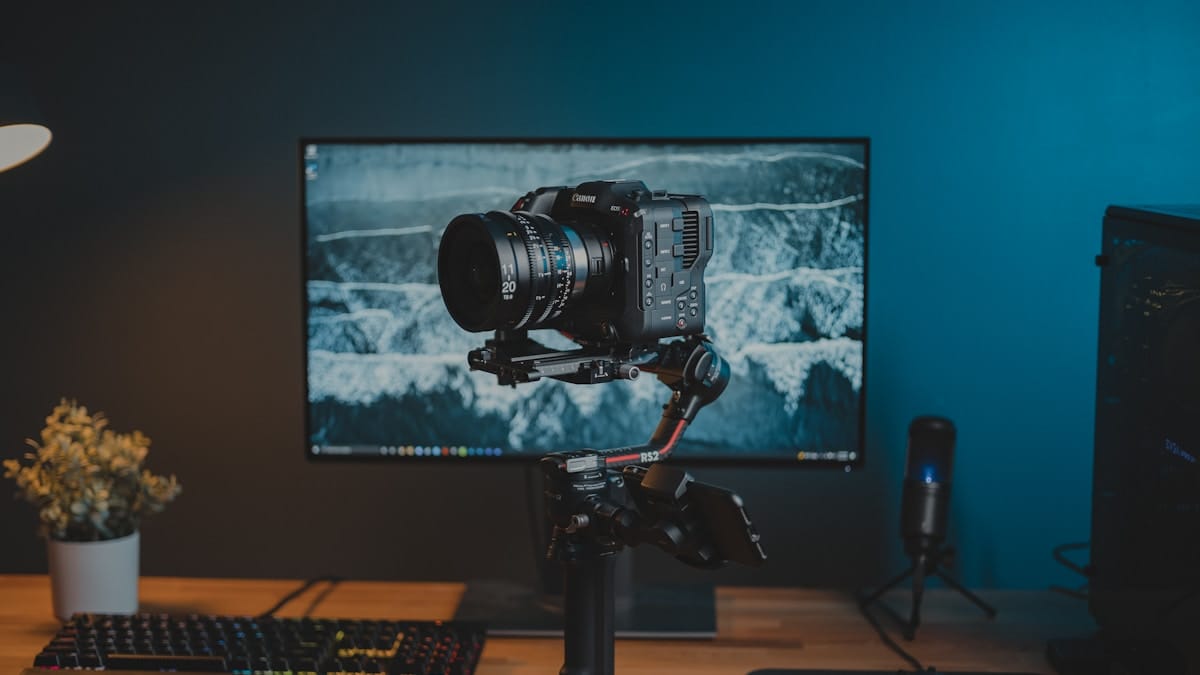 A digital camera mounted on a tripod is positioned in front of a computer monitor on a desk setup.