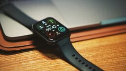A black smartwatch with a digital display showing 09:42 lies on a wooden surface next to a closed laptop.