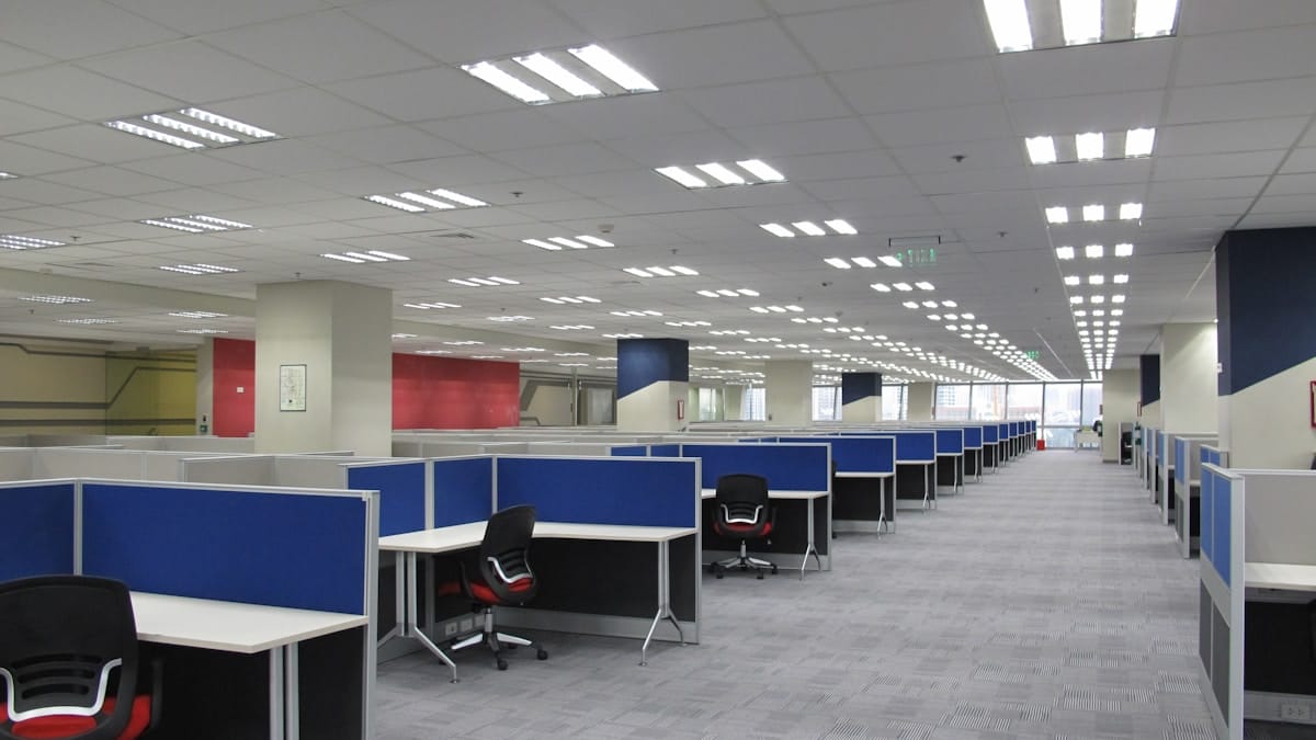 A modern open-plan office with rows of empty cubicles, blue dividers, and black chairs under bright ceiling lights.