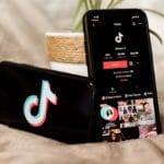 Two smartphones display the TikTok logo and TikTok app profile page, resting on a blanket next to a small potted plant.