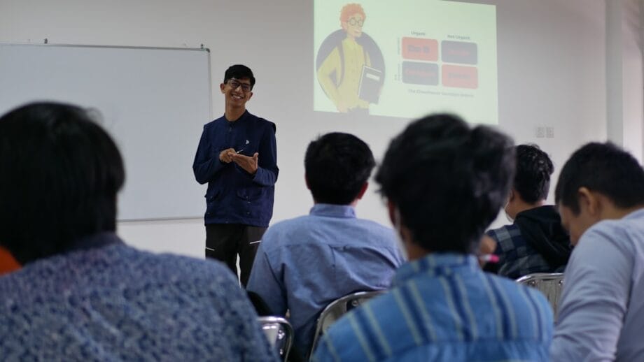 A person stands at the front of a classroom giving a presentation to seated students, with a slide projected on the wall.