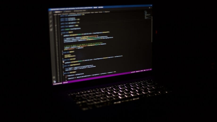 A dimly lit laptop displays lines of colorful code on the screen against a dark background.