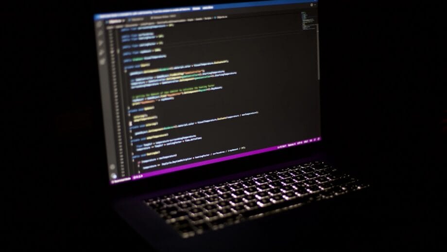 A laptop in a dark room displays code on its screen in a text editor with a black background.