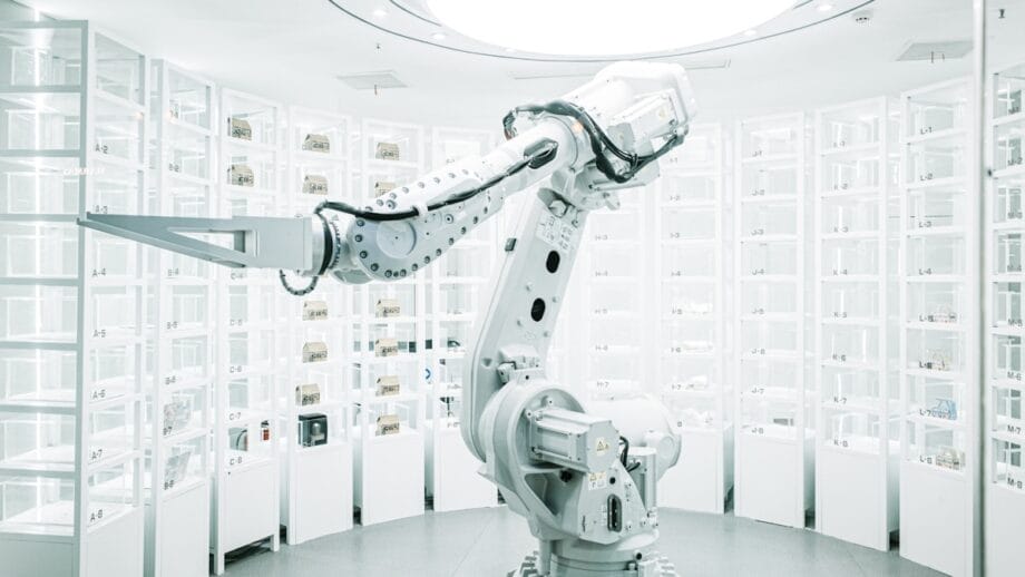 A robotic arm operates in a bright, white laboratory with shelves filled with various small items or containers.