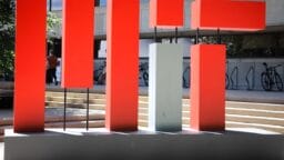 Large red and gray abstract sculpture spelling MIT stands outside a building with bike racks and steps in the background.