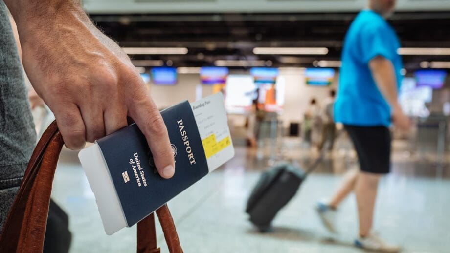 A person holds a U.S. passport and boarding pass in an airport terminal with travelers and check-in counters in the background.