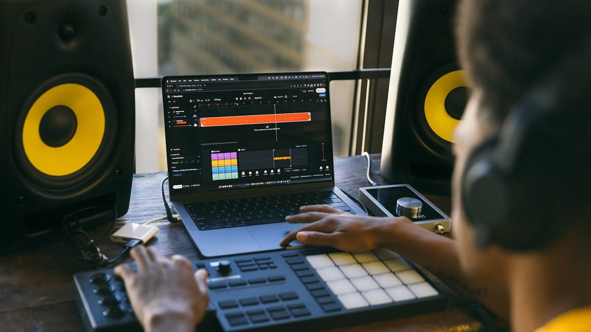 Person using a laptop and MIDI controller to produce music, with headphones on and studio speakers positioned on each side.