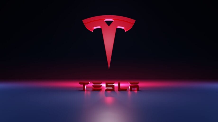Red Tesla logo above the word TESLA in bold 3D letters, set against a dark background with pink and blue lighting effects.