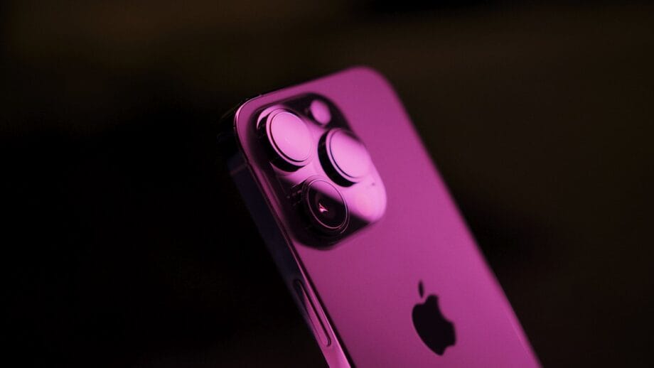 Close-up of the back of a smartphone with three camera lenses, lit by pink light, showing part of the Apple logo.