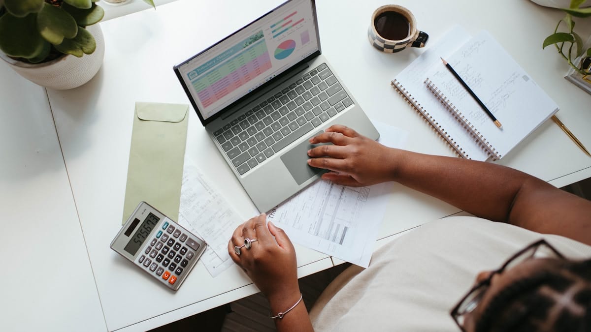 Person working at a desk with a laptop displaying charts, a calculator, documents, a notepad, and a cup of coffee.
