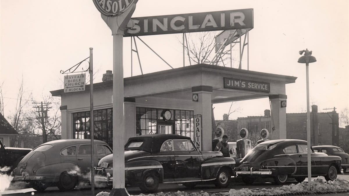 Black-and-white photo of three vintage cars at a Sinclair gas station called Jim’s Service, with visible gas pumps and signage.