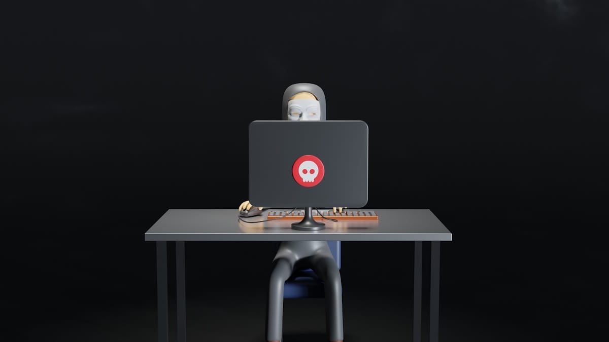 3D cartoon figure sits at a desk using a computer with a red skull icon on the monitor against a dark background.
