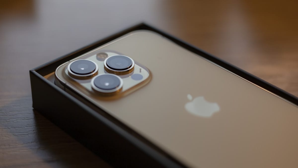 A gold smartphone with three camera lenses, partially removed from its black box, sits on a wooden surface.