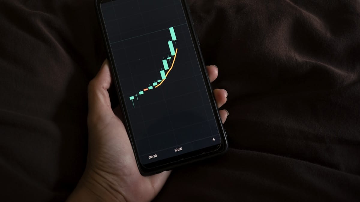 A hand holds a smartphone displaying a candlestick chart with a sharp upward trend on the screen.