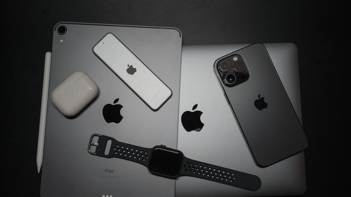 Assorted Apple devices including an iPad, MacBook, iPhone, Apple Watch, AirPods, Apple Pencil, and remote on a black surface.