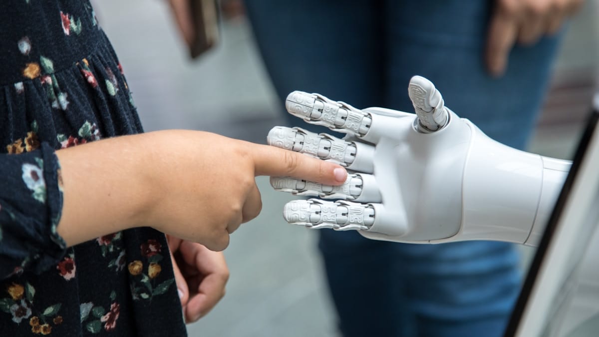 A child touches the finger of a humanoid robot hand, with an adult standing in the background.