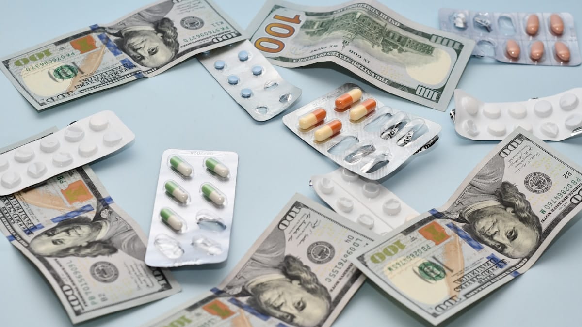 Blister packs of pills and capsules are scattered among several US 100-dollar bills on a light blue surface.
