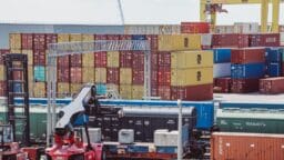 Stacks of colorful shipping containers and freight cars at a busy industrial port with cranes and equipment in the background.
