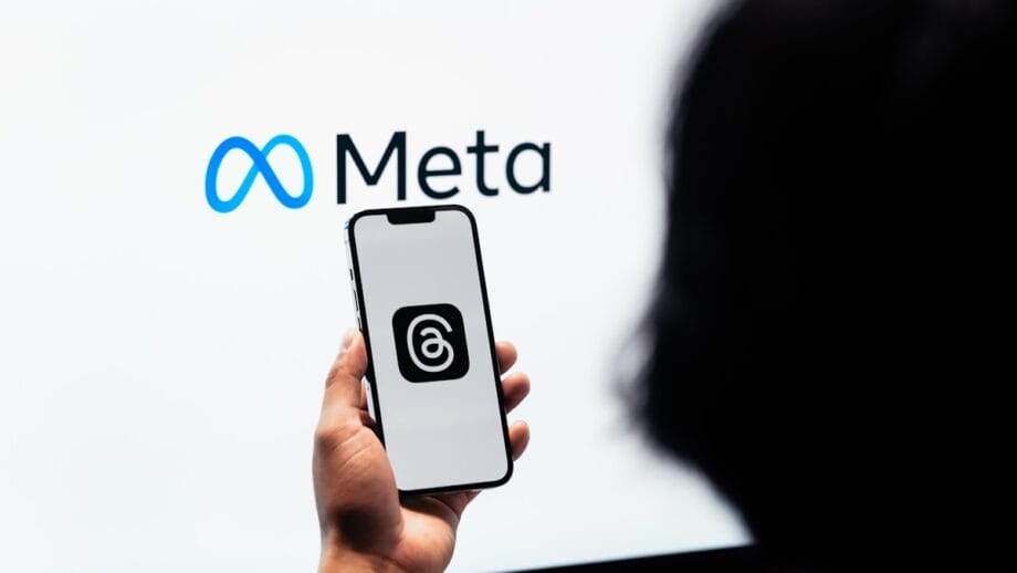 A person holds a smartphone displaying the Threads app logo, with the Meta logo visible on a screen in the background.