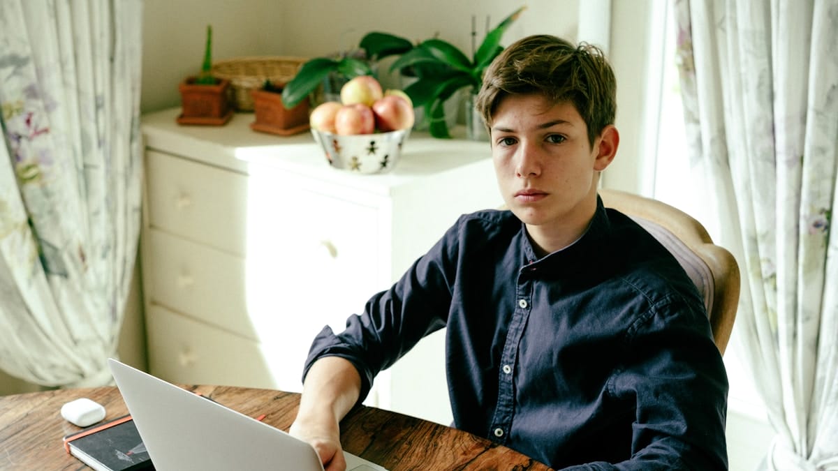 Teen boy sits at a table using a laptop, with fruit and plants in the background, in a well-lit room with floral curtains.