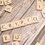 Scrabble tiles spelling “CRYPTO” are arranged on a wooden surface, surrounded by scattered letter tiles.
