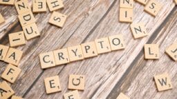 Scrabble tiles spelling “CRYPTO” are arranged on a wooden surface, surrounded by scattered letter tiles.