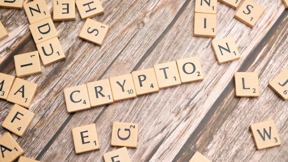 Scrabble tiles spelling “CRYPTO” are arranged on a wooden surface, surrounded by scattered letter tiles.