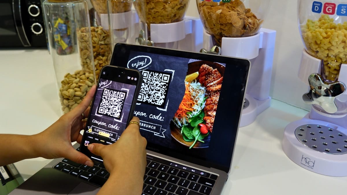 Person scans a QR code for a coupon displayed on a laptop screen showing a plated meal, with cereal dispensers in the background.