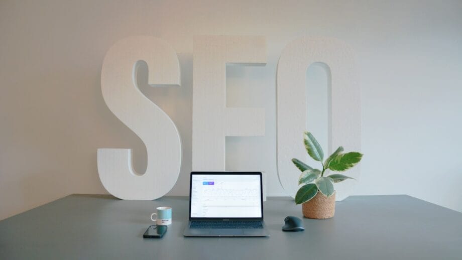 A laptop, phone, mug, and potted plant on a desk with large white letters spelling SEO in the background.