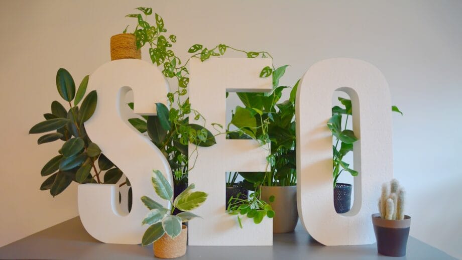Large white letters spelling SEO are placed in front of several potted green plants on a gray table against a plain wall.