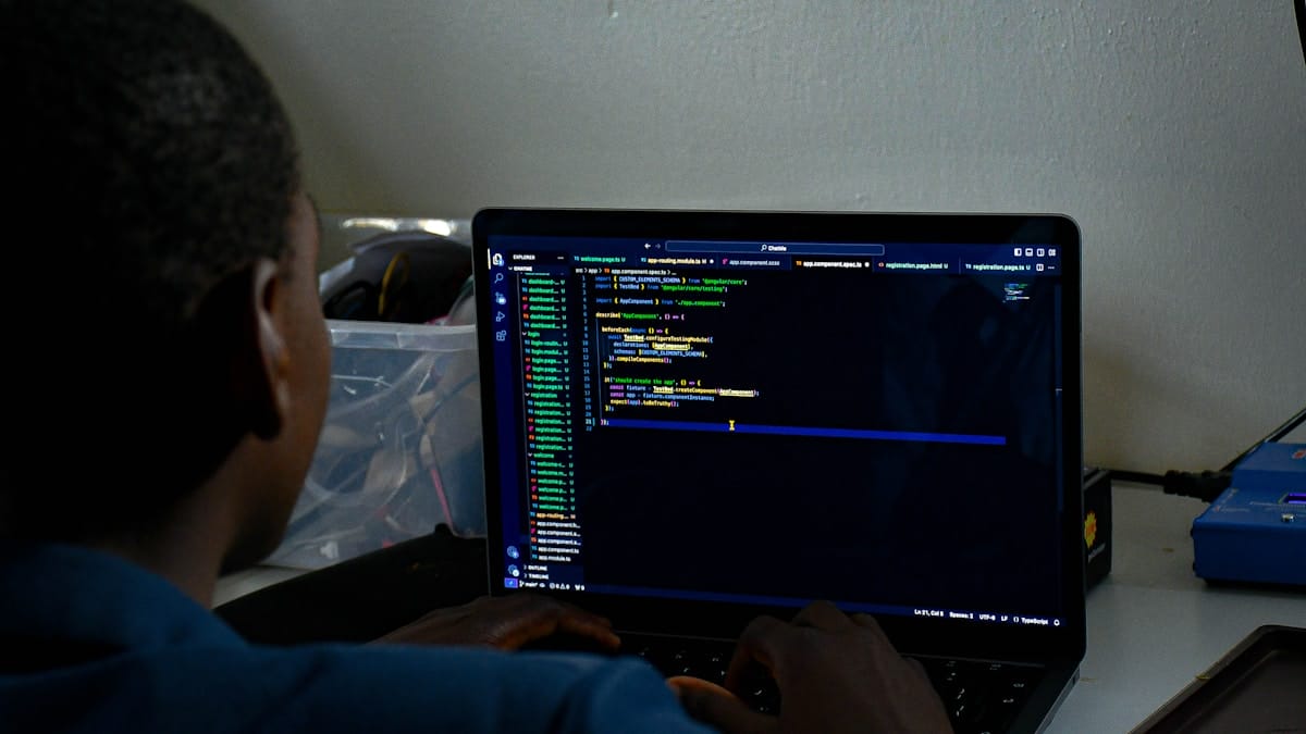 A person sits at a desk typing code on a laptop displaying a dark-themed programming interface.