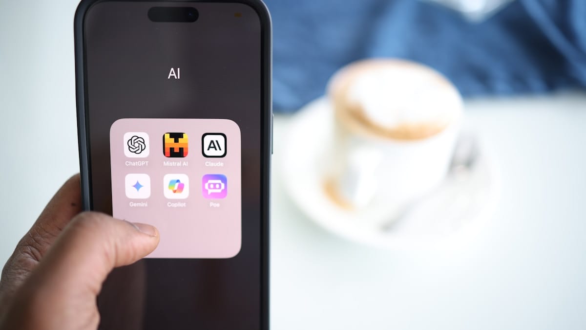 A hand holds a smartphone displaying a folder labeled AI with various AI app icons; a cup sits in the blurred background.