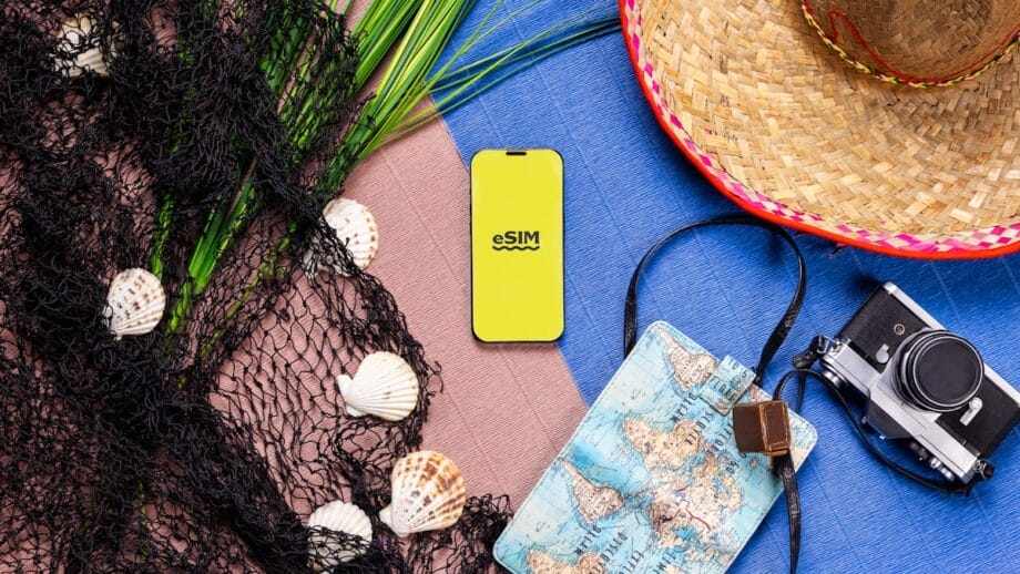 Smartphone with eSIM on screen, seashells, fishing net, straw hat, map, and camera arranged on colorful background.