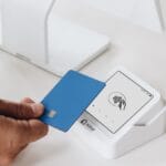 A person holds a blue credit card near a white contactless payment terminal on a white counter.