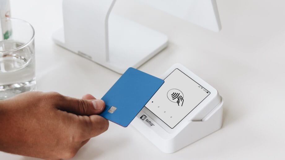 A person holds a blue credit card near a white contactless payment terminal on a white counter.