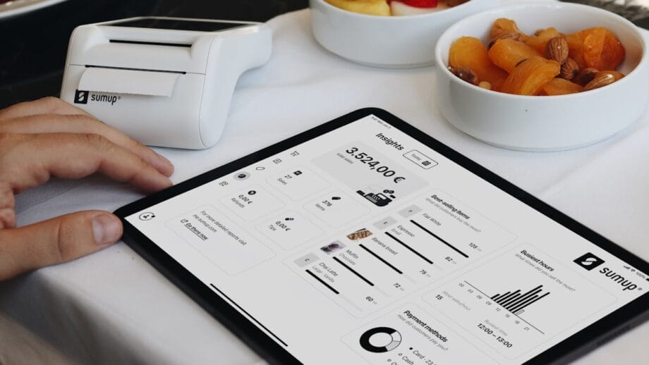 A person uses a tablet showing financial analytics and graphs, with a portable receipt printer and a bowl of dried fruit nearby on a table.