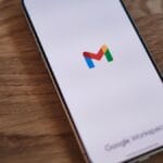 A smartphone displays the Gmail app loading screen, featuring the Gmail logo, on a wooden surface.