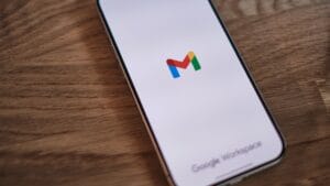 A smartphone displays the Gmail app loading screen, featuring the Gmail logo, on a wooden surface.