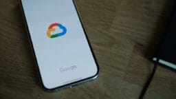 A smartphone displaying the Google Cloud logo on its screen rests on a wooden surface next to a notebook.