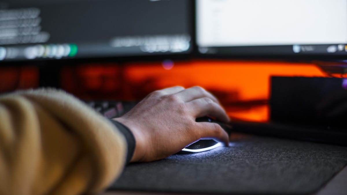 A person’s hand using a computer mouse on a mouse pad, in front of two monitors with a red-orange light in the background.