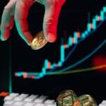 A hand holds a Bitcoin coin near a keyboard, with a cryptocurrency price chart displayed on a screen in the background.