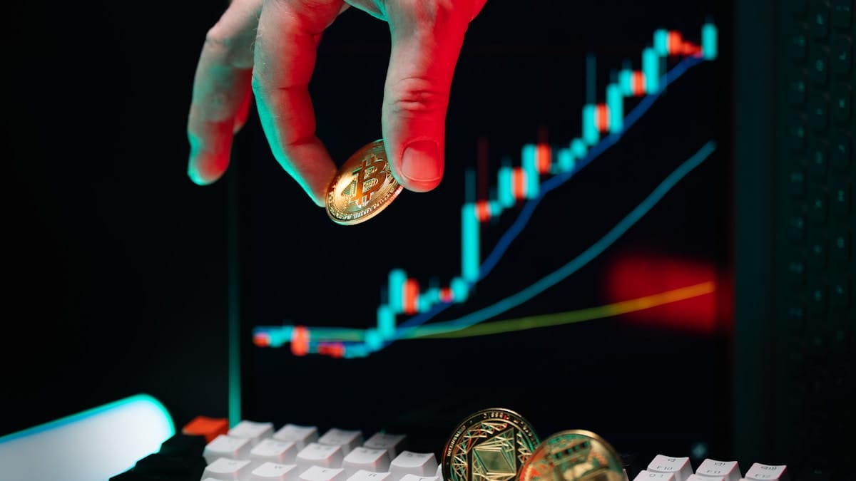 A hand holds a Bitcoin coin near a keyboard, with a cryptocurrency price chart displayed on a screen in the background.