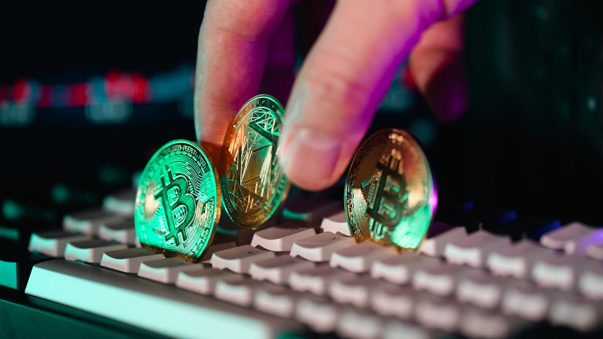 A hand holds three cryptocurrency coins—Bitcoin, Ethereum, and another—above a computer keyboard.