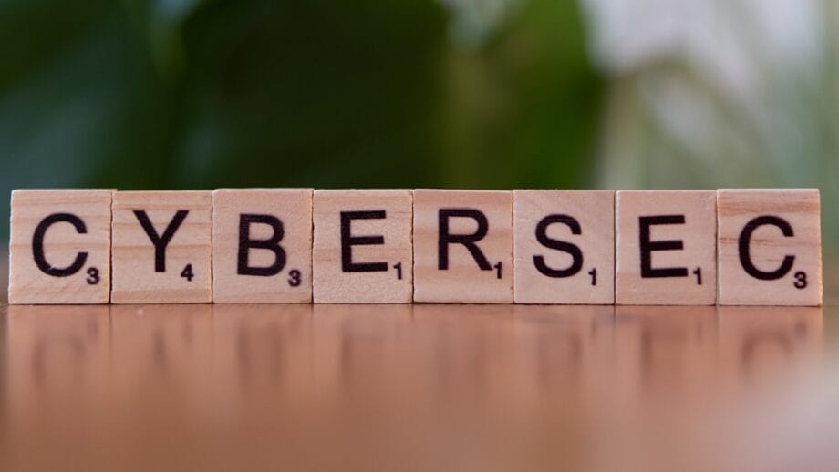 Seven wooden letter tiles spell out CYBERSEC on a wooden surface with a green blurred background.