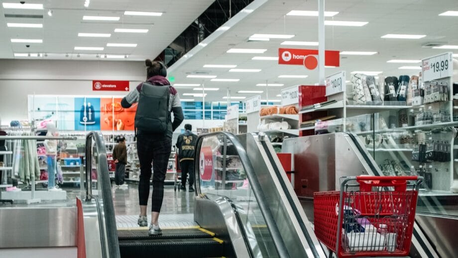 A person with a backpack rides an escalator inside a retail store, with shopping carts and merchandise visible.