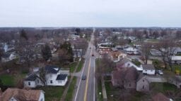 A straight road runs through a small town with houses, trees, and cars on an overcast day.