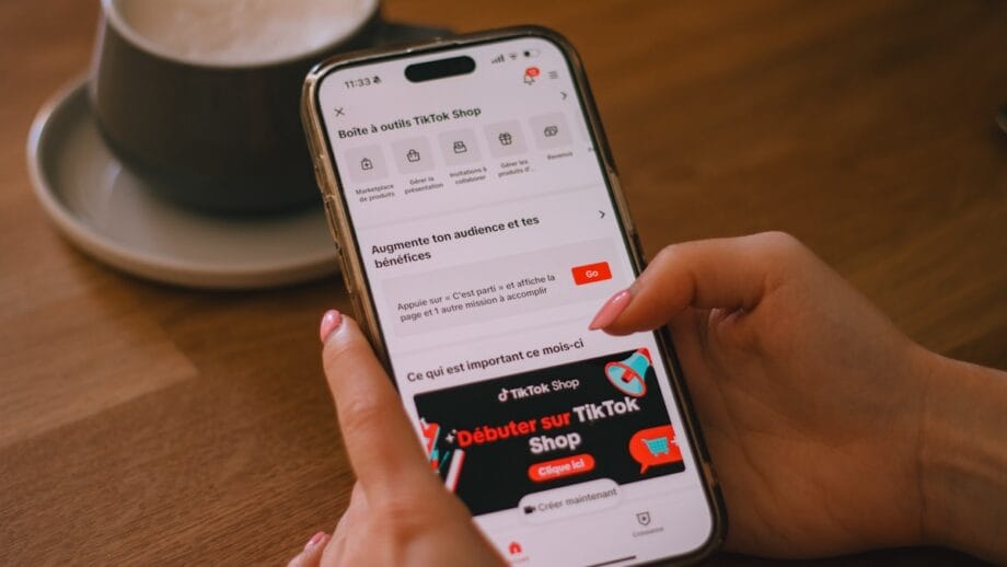 Person holding a smartphone displaying the TikTok Shop interface, with a cup and saucer in the background on a wooden table.