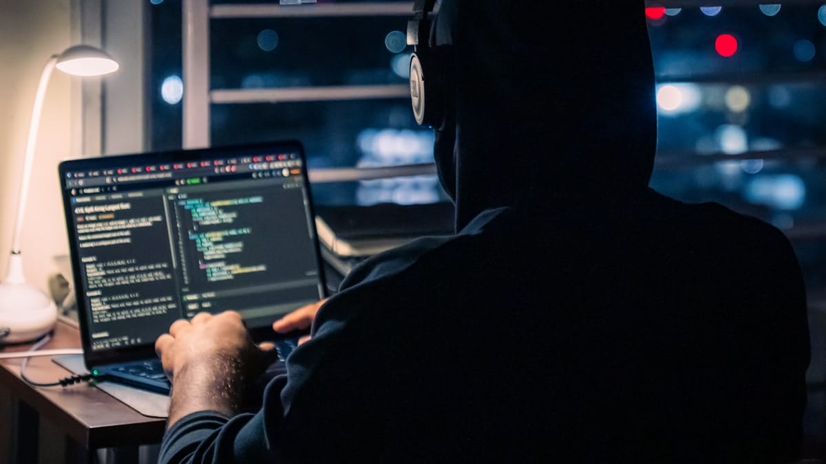 Person wearing a hoodie and headphones working on a laptop with code on the screen at a desk near a window at night.