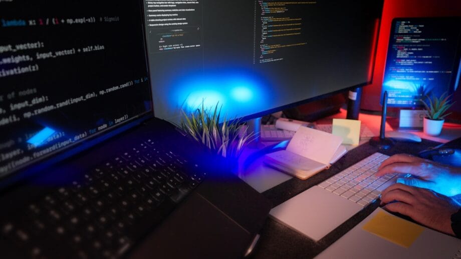 A person types on a keyboard at a desk with multiple monitors displaying code, a laptop, and small potted plants.